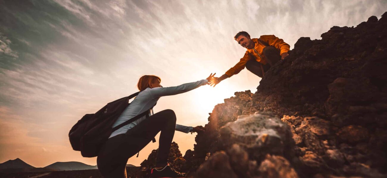 Couple,Of,Hikers,Helping,Each,Other,Climbing,A,Mountain,At